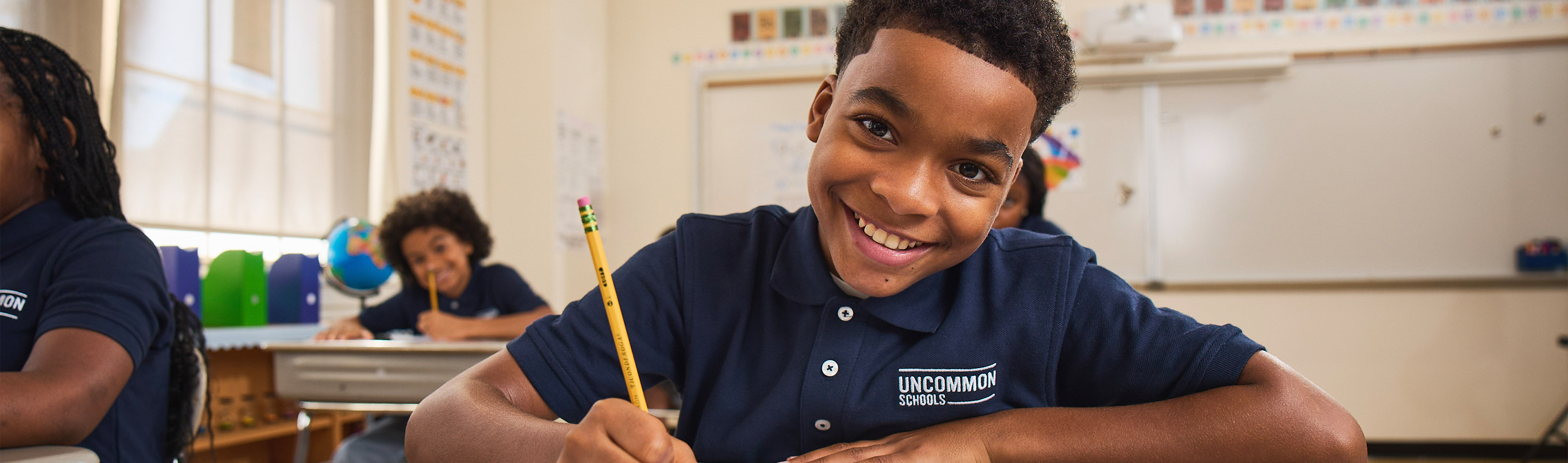 Smiling Uncommon Schools student holding a pencil at his desk in a bright classroom