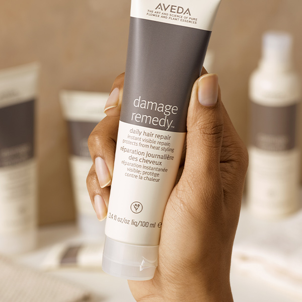 Hand holding an Aveda Damage Remedy daily hair repair tube with other products softly blurred in the background, campaign content by The Charles Group