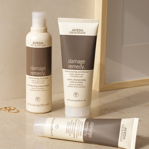 Aveda Damage Remedy restructuring shampoo, conditioner, and daily hair repair tube arranged on a warm beige surface, campaign content by The Charles Group