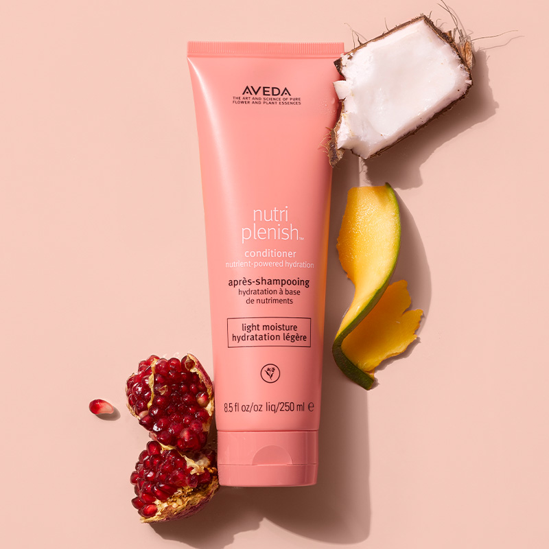 Aveda Nutriplenish light moisture conditioner tube surrounded by pomegranate, coconut, and mango ingredients on a peach background