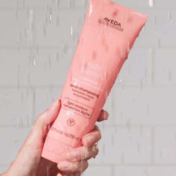 Hand holding Aveda Nutriplenish conditioner tube in a shower with water droplets falling around it
