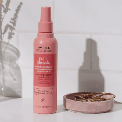 Aveda Nutriplenish leave-in conditioner spray bottle styled on a bathroom surface next to a pink dish and hair accessories