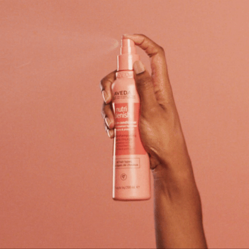 Hand holding and spraying Aveda Nutriplenish leave-in conditioner against a warm peach background