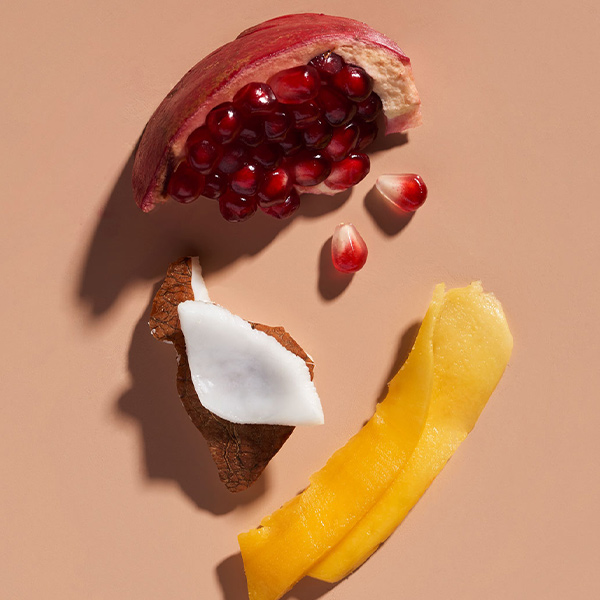 Styled flatlay of Aveda Nutriplenish key ingredients including pomegranate seeds, coconut, and mango on a warm peach background, campaign content by The Charles Group