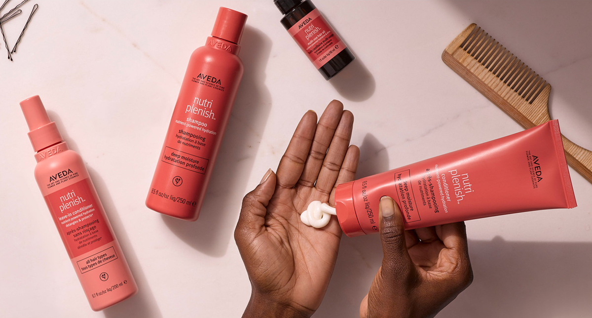 Model's hands applying Aveda Nutriplenish conditioner from tube, surrounded by full product collection and wooden comb