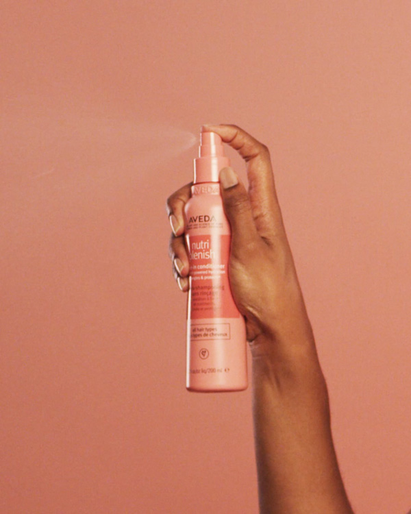 Hand holding an Aveda Nutriplenish leave-in conditioner spray bottle mid-spritz against a coral-pink background, social video still produced by The Charles Group