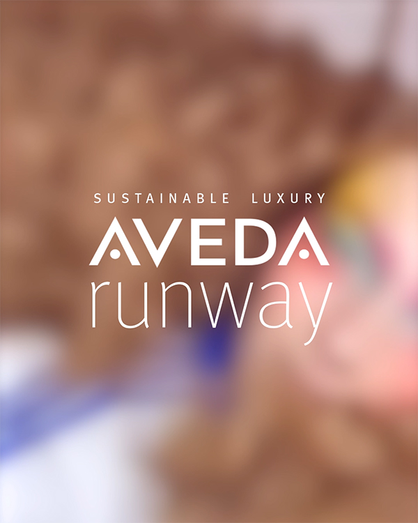 Aveda Runway 'Sustainable Luxury' event title card with blurred backstage fashion models in the background, live event content by The Charles Group