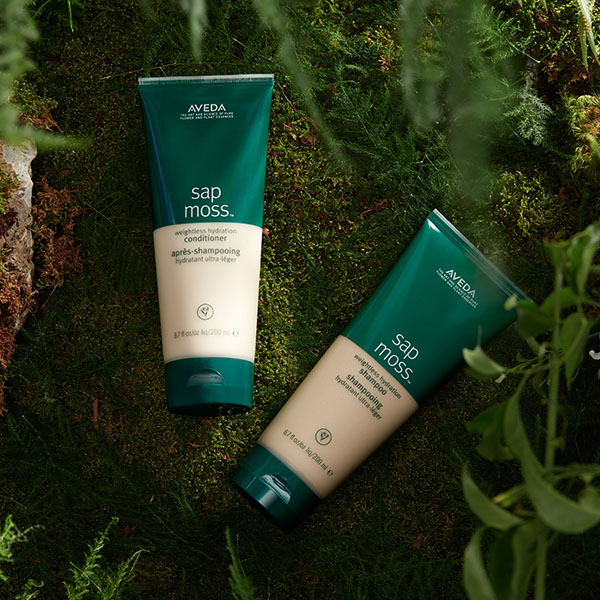 Aveda Sap Moss shampoo and conditioner bottles against a dark mossy natural background