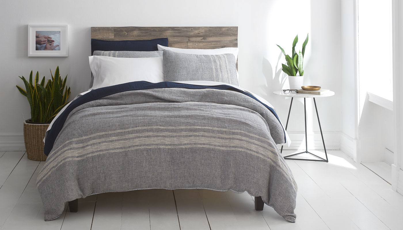 Threaded Home linen bedding campaign hero — styled bed with grey stripe coverlet and navy accents, produced by The Charles Group