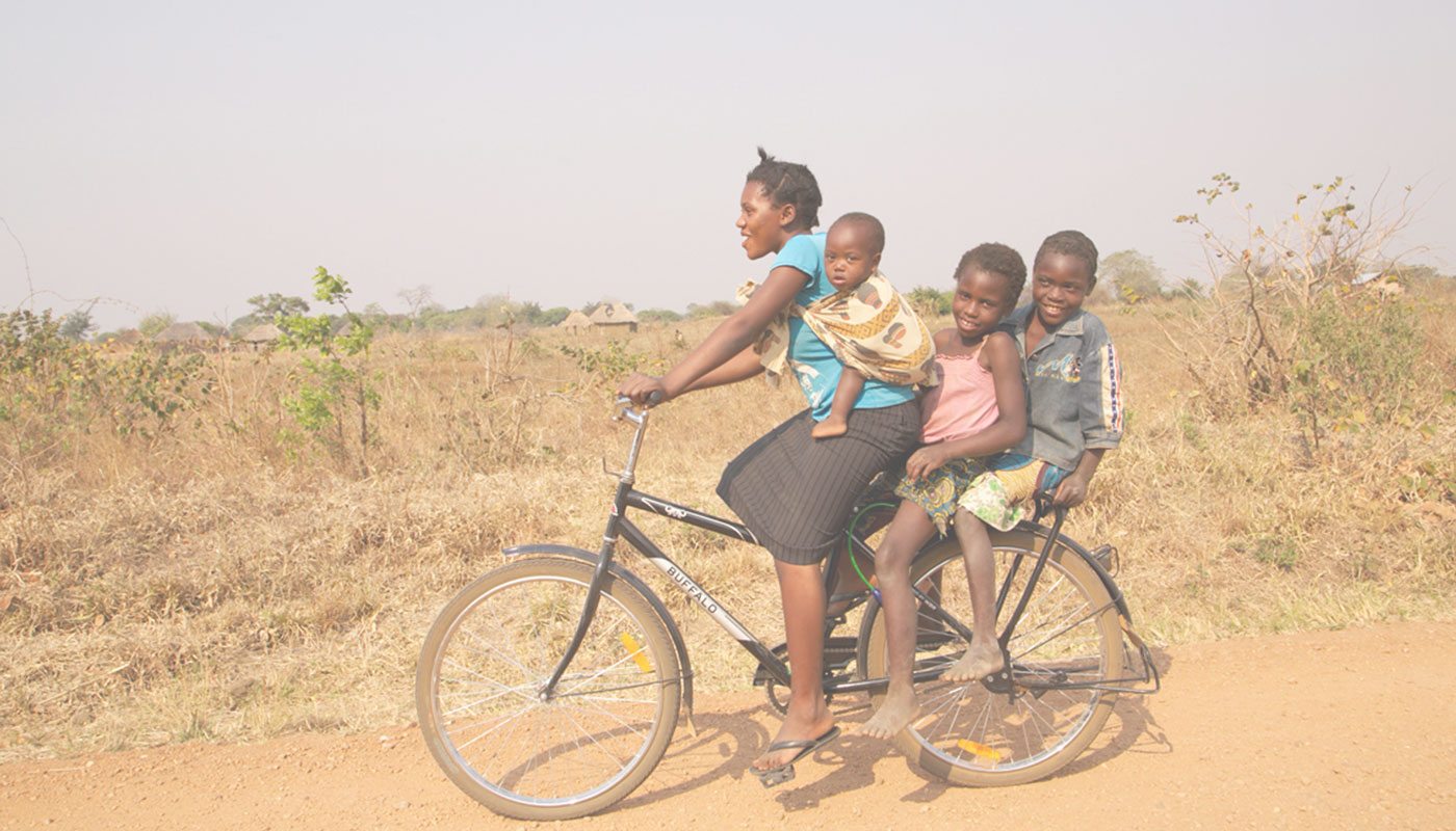 A smiling mother carries three children on a Buffalo Bicycle along a rural African dirt road — World Bicycle Relief campaign photography by The Charles Group