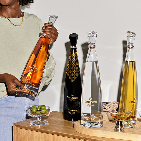 Woman selecting a Cincoro tequila bottle from a lineup of four distinct expressions displayed on a wooden sideboard