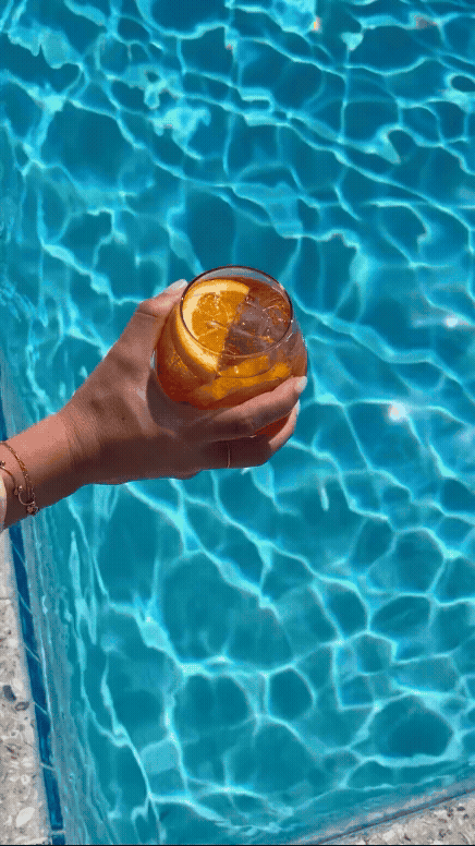 Hand holding an iced cocktail with an orange slice above a sparkling turquoise pool at The Seagate Delray